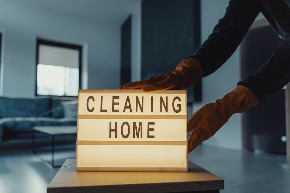 A close-up of a person wearing dark clothing and brown cleaning gloves, gently adjusting a lightbox sign that reads 'CLEANING HOME' on a wooden surface in a modern, minimalist living room with a sofa,