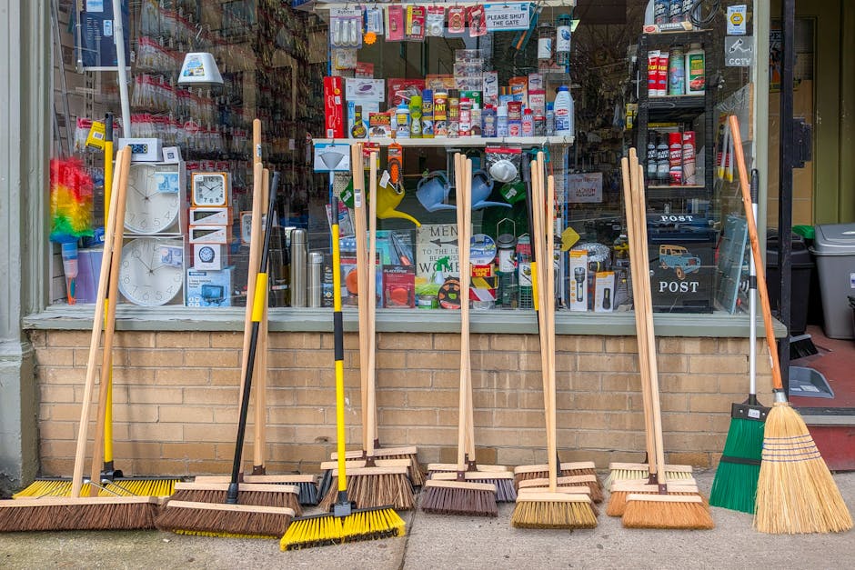 Exterior view of a retail shop on Kilburn High Road showcasing a variety of cleaning tools and supplies on the sidewalk and within the store. Visible items include brooms, mops, buckets, plastic stora